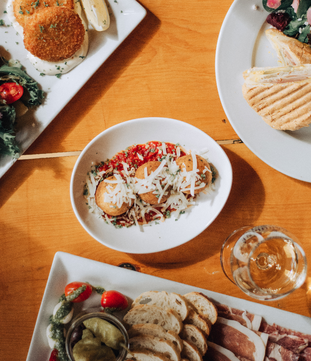 Overhead view of a wooden table set with assorted dishes from Caroline Cellars Winery. In the center, a white bowl features arancini topped with marinara sauce, grated cheese, and herbs. Surrounding it are plates with a charcuterie board including sliced bread, cured meats, and pickled vegetables; a panini sandwich with a side salad; and crispy croquettes served with dip. A glass of white wine sits nearby, complementing the rustic, winery-style spread.